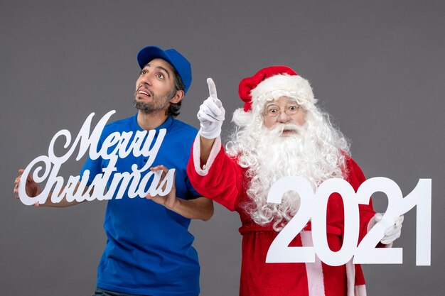 Front view of santa claus with male courier holding merry christmas and 2021 banner on a grey wall