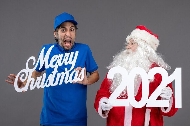 Front view of santa claus with male courier holding merry christmas and 2021 boards on a grey wall