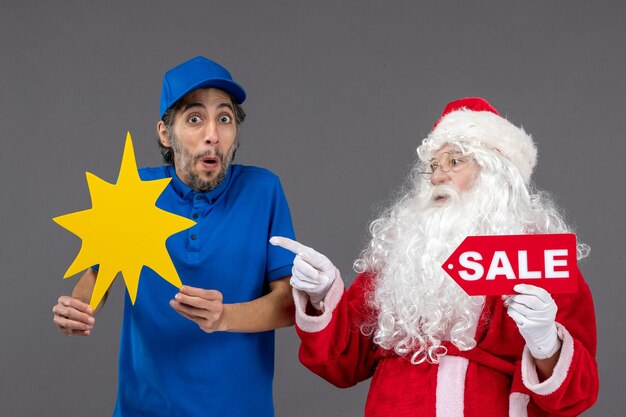 Front view of santa claus with male courier holding sale banner and yellow sign on a grey wall