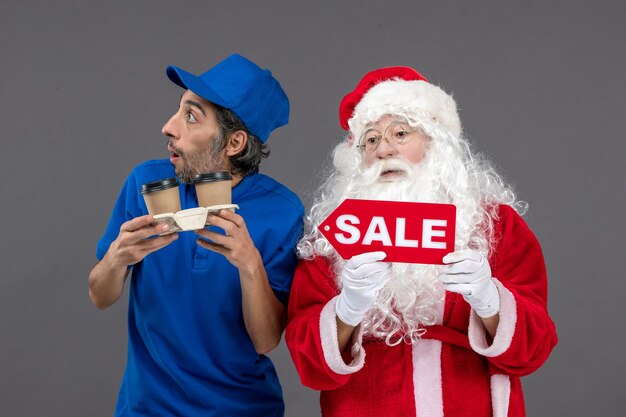 Front view of santa claus with male courier holding sale board and coffee on the grey wall