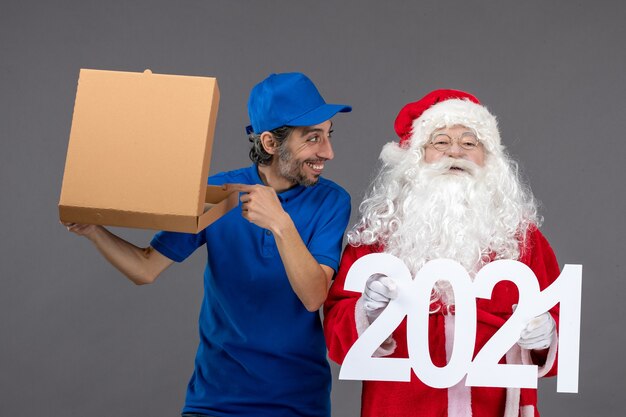 Front view of santa claus with male courier holding shopping bags and food box on grey wall