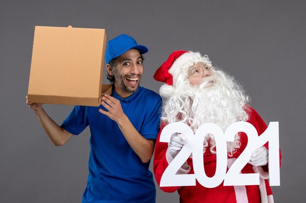 Front view of santa claus with male courier holding shopping bags and food box on the grey wall