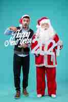 Free photo front view of santa claus with male holding happy new year and 2021 boards on a blue wall