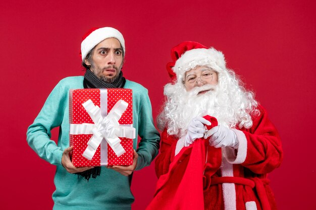 Вид спереди санта-клауса с мужчиной, вынимающим подарок из сумки на красном столе, цвет рождественской эмоции красный