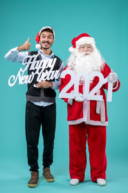 Free photo front view of santa claus with man holding happy new year and 2021 boards on blue wall