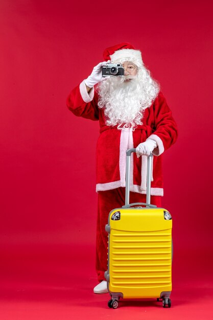 Front view of santa claus with yellow bag taking photo with camera on a red wall