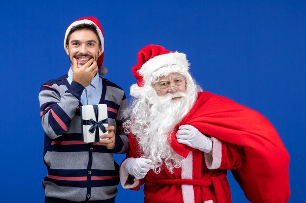 Front view santa claus with young male carrying bag full of presents on blue color holiday presents xmas