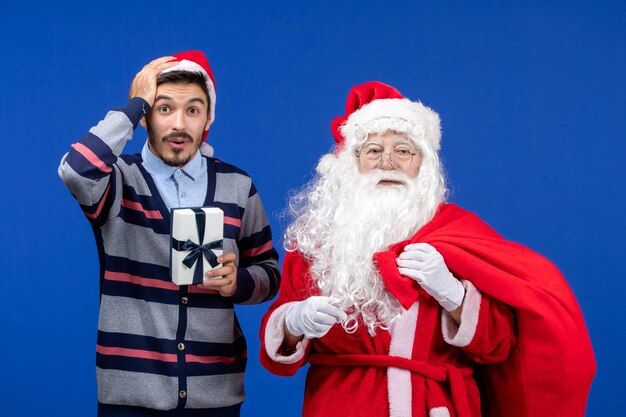 青い感情の休日のプレゼントクリスマスにプレゼントでいっぱいの若い男性のキャリーバッグと正面のサンタクロース