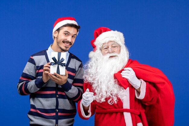 Front view santa claus with young male carrying bag full of presents on blue floor color holiday