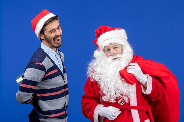 青いプレゼントクリスマスの休日の色のプレゼントでいっぱいの若い男性のキャリングバッグと正面図のサンタクロース
