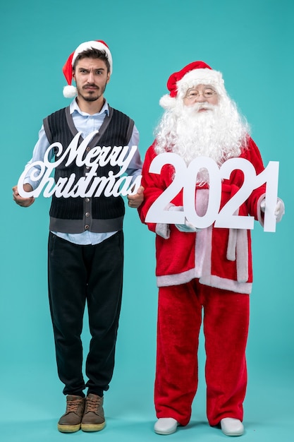 Free photo front view santa claus with young male holding merry christmas and writings on blue desk