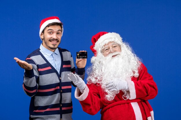 Front view of santa claus with young man holding bank card on a blue wall