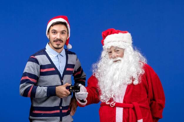 파란색 벽에 은행 카드를 들고 있는 젊은 남자와 산타 클로스의 전면 보기