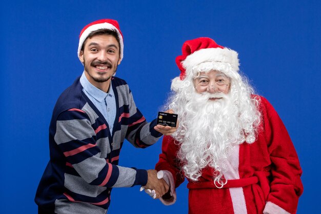 Front view of santa claus with young man holding bank card on blue wall