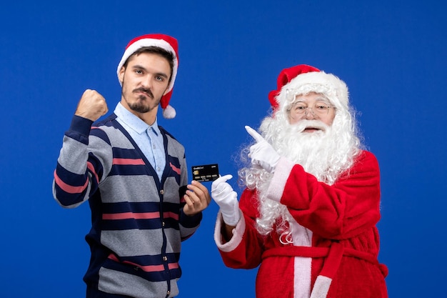 Front view of santa claus with young man holding bank card on blue wall