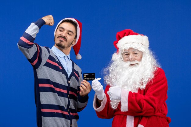 Front view of santa claus with young man holding bank card on blue wall