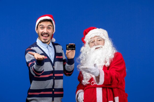 Front view of santa claus with young man holding bank card on the blue wall