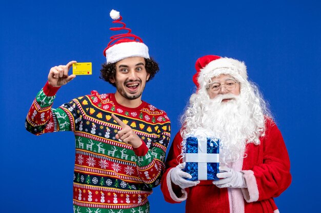 은행 카드를 들고 파란색 벽에 있는 젊은 남자와 산타 클로스의 전면 보기