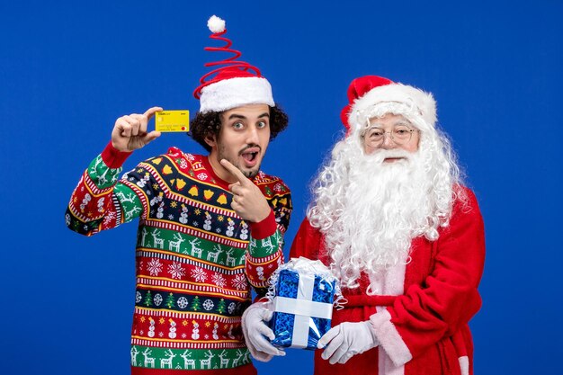 Front view of santa claus with young man holding bank card and present on blue wall