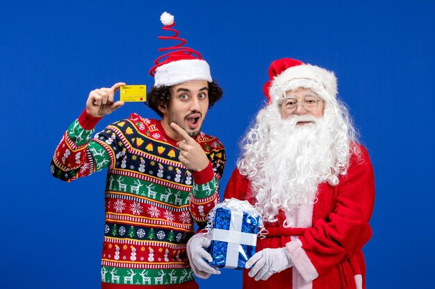 은행 카드를 들고 파란색 벽에 있는 젊은 남자와 산타 클로스의 전면 보기