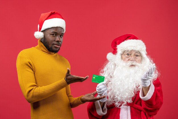 Free photo front view of santa claus with young man holding bank card on a red wall