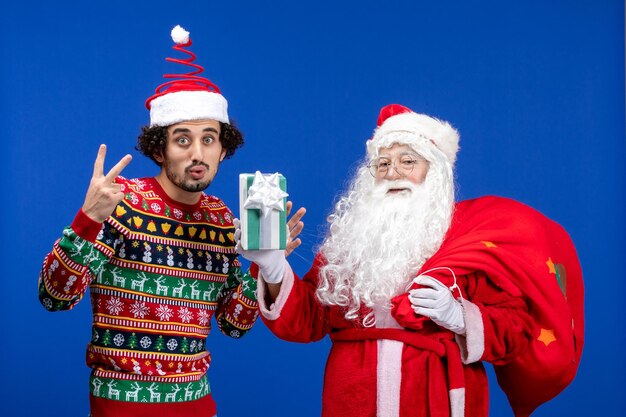 Front view of santa claus with young man holding christmas present on a blue wall