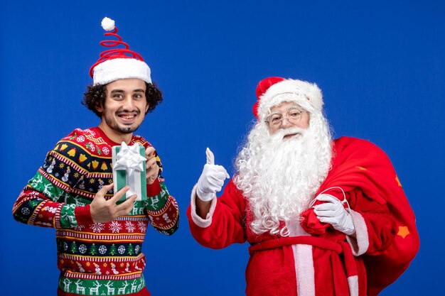 Front view of santa claus with young man holding christmas present on blue wall