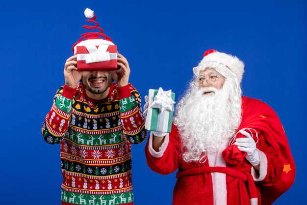 Front view of santa claus with young man holding christmas presents on a blue wall