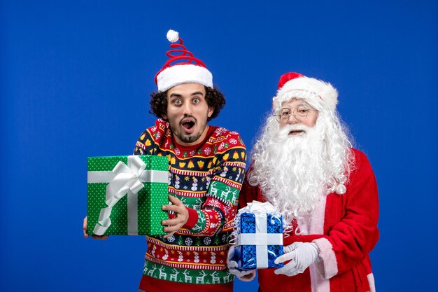 파란색 벽에 크리스마스 선물을 들고 있는 젊은 남자와 산타 클로스의 전면 보기
