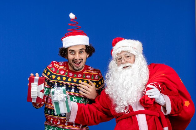 Front view of santa claus with young man holding christmas presents on blue wall