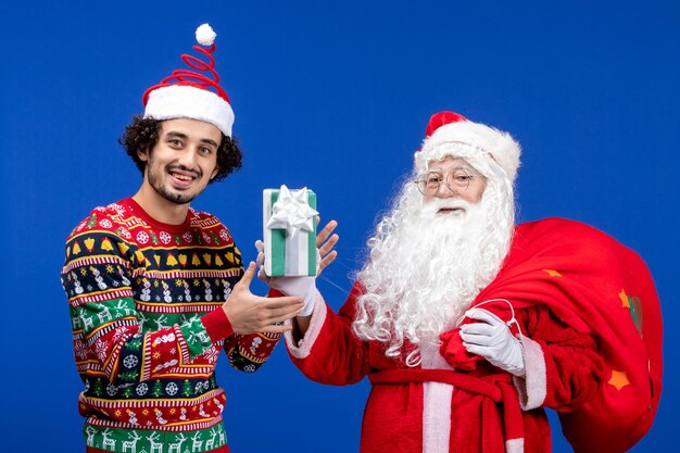 Front view of santa claus with young man holding christmas presents on blue wall