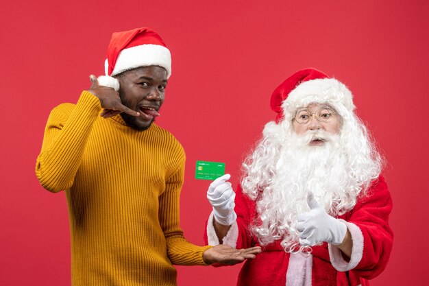 Front view of santa claus with young man holding green bank card on red wall