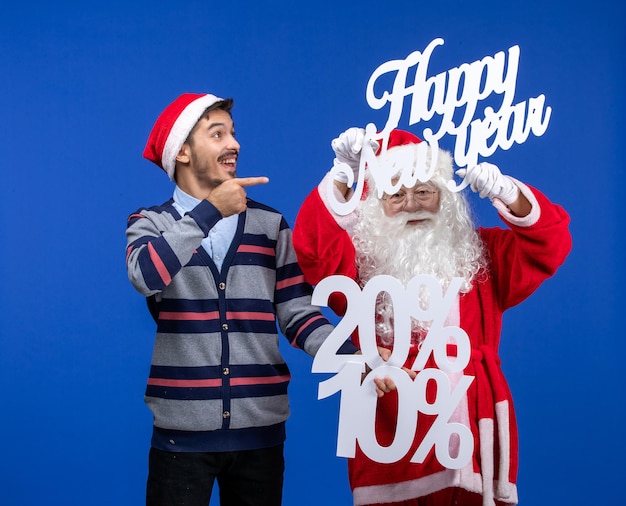 Foto gratuita vista frontale di babbo natale con un giovane che tiene in mano felice anno nuovo e numeri scritti sul muro blu