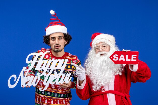 Front view of santa claus with young man holding happy New Year's and sale writings on blue wall