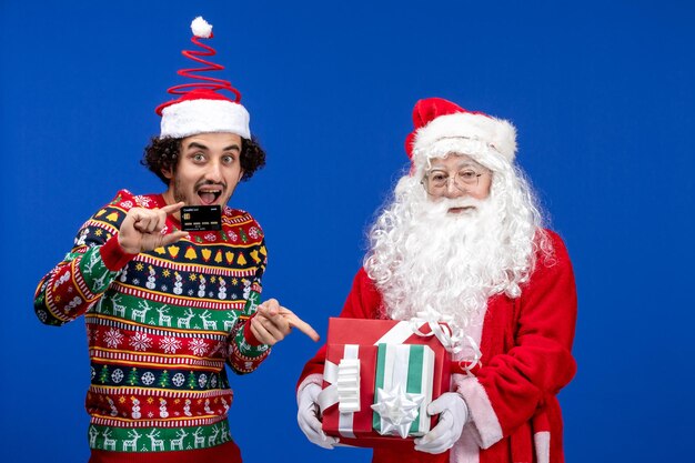 Front view of santa claus with young man holding presents and bank card on blue wall