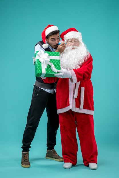 Free photo front view of santa claus with young man and holiday present on blue wall