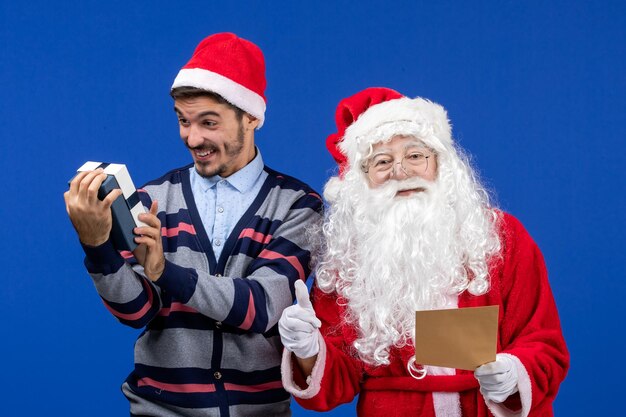 Front view of santa claus with young man reading letter and holding present on blue wall