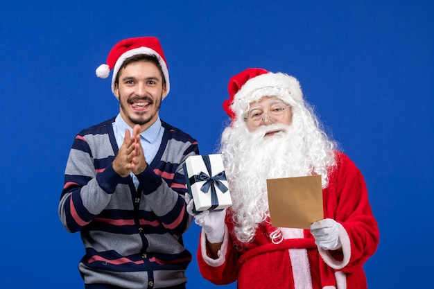 Front view of santa claus with young man reading letter and holding present on blue wall