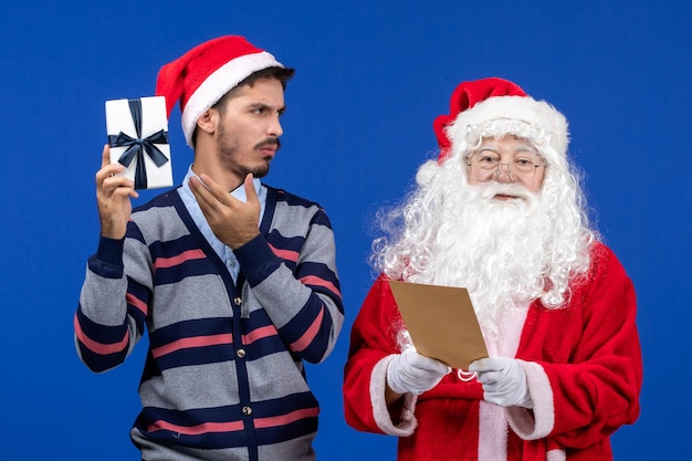 Front view of santa claus with young man reading letter and holding present on blue wall