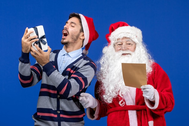 Front view of santa claus with young man reading letter and holding present on blue wall