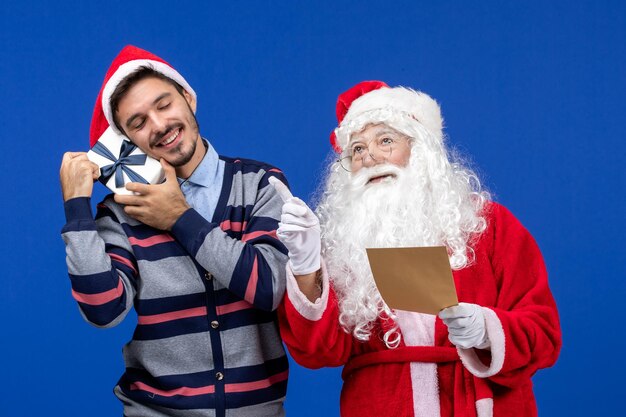 Front view of santa claus with young man reading letter and holding present on blue wall