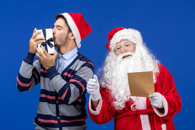 Front view of santa claus with young man reading letter and holding present on the blue wall