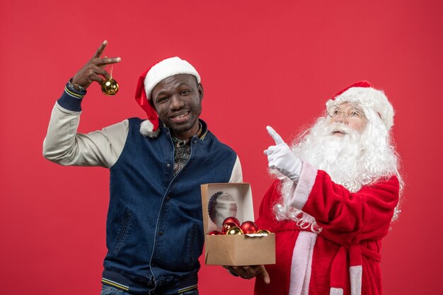 Front view of santa claus with young young man holding xmas tree toys on red wall