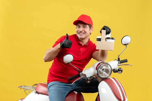 Front view of satisfied confident courier man wearing red blouse and hat gloves in medical mask delivering order sitting on scooter holding orders making ok gesture