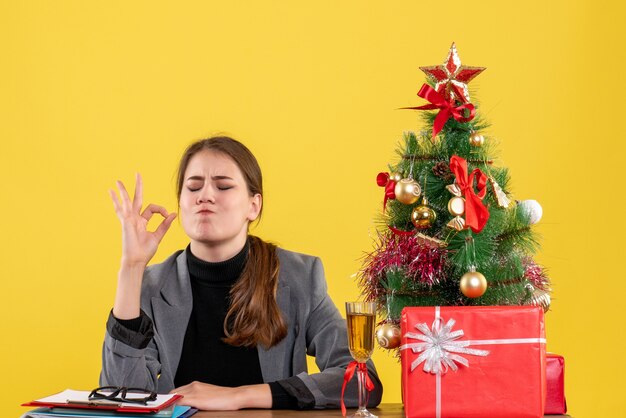 Front view satisfied girl sitting at the desk making okey sign near xmas tree and gifts cocktail