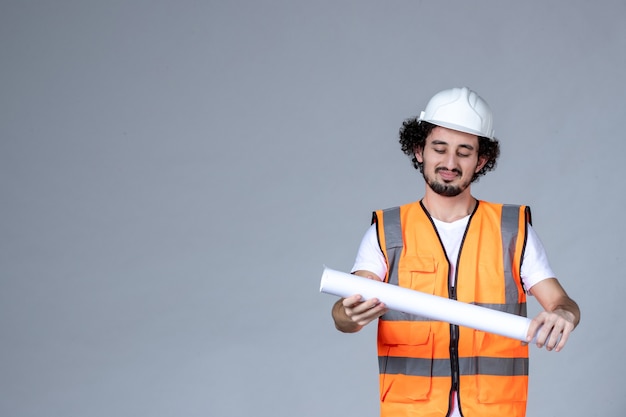 Front view of satisfied male constructor in warning vest wearing safety helmet and holding blank on gray wave wall