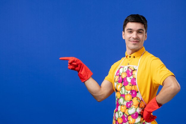 Front view of satisfied male housekeeper in apron putting hand on a waist standing on blue wall