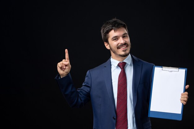 Front view of satisfied young adult in suit holding free space for writing and pointing up on isolated dark wall