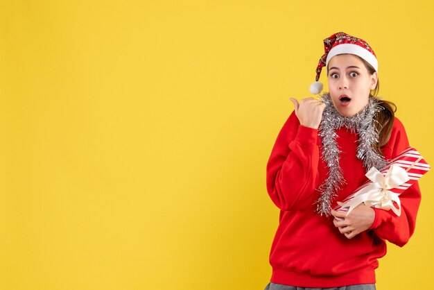 Front view scared girl with red sweater and santa hat holding her gift showing something