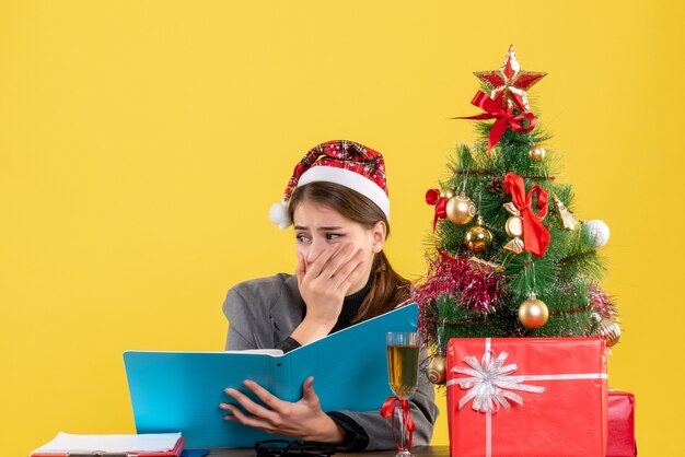 Front view scared girl with xmas hat sitting at the table holding documents xmas tree and gifts cocktail